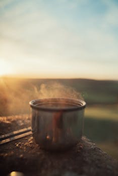 Close-up of a steaming hot drink in a metal cup with sunrise in the background.