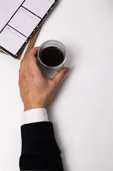 Close-up of a hand holding a coffee cup on a white desk with a notepad visible.