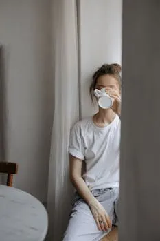 Calm young woman in a white t-shirt sipping coffee by a window indoors.
