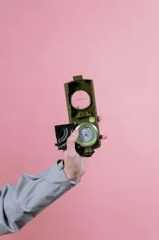 A hand holding a military compass against a pastel pink background,