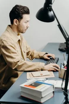 A man in a brown jacket working at a desk in an office setting, typing on a