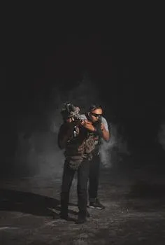 Two men in combat gear simulate a tactical operation amid smoke and darkness.