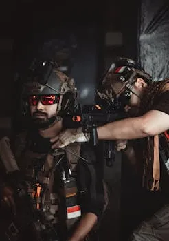 Two soldiers in full tactical gear aiming a rifle in a dimly lit setting.