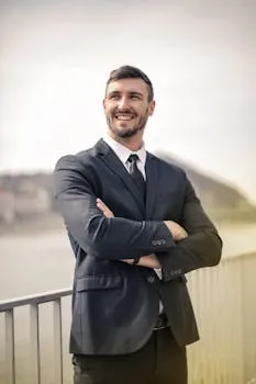 Smiling businessman in a black suit standing outdoors by a railing,