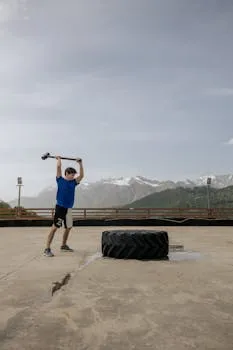 A man swings a sledgehammer to train with a tire in a scenic outdoor setting.